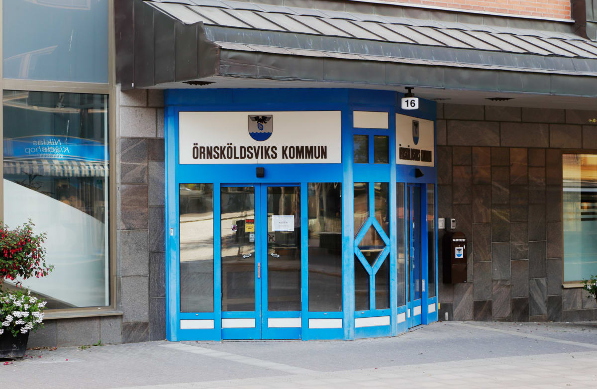 En blå- och vitmålad entré till en byggnad med texten "ÖRNSKÖLDSVIKS KOMMUN" och ett kommunvapen ovanför dörrarna. Entrén har fönster och en siffra "16" ovanför. Intill finns en fönsterruta som reflekterar omgivningen och en växtkruka med blommor.