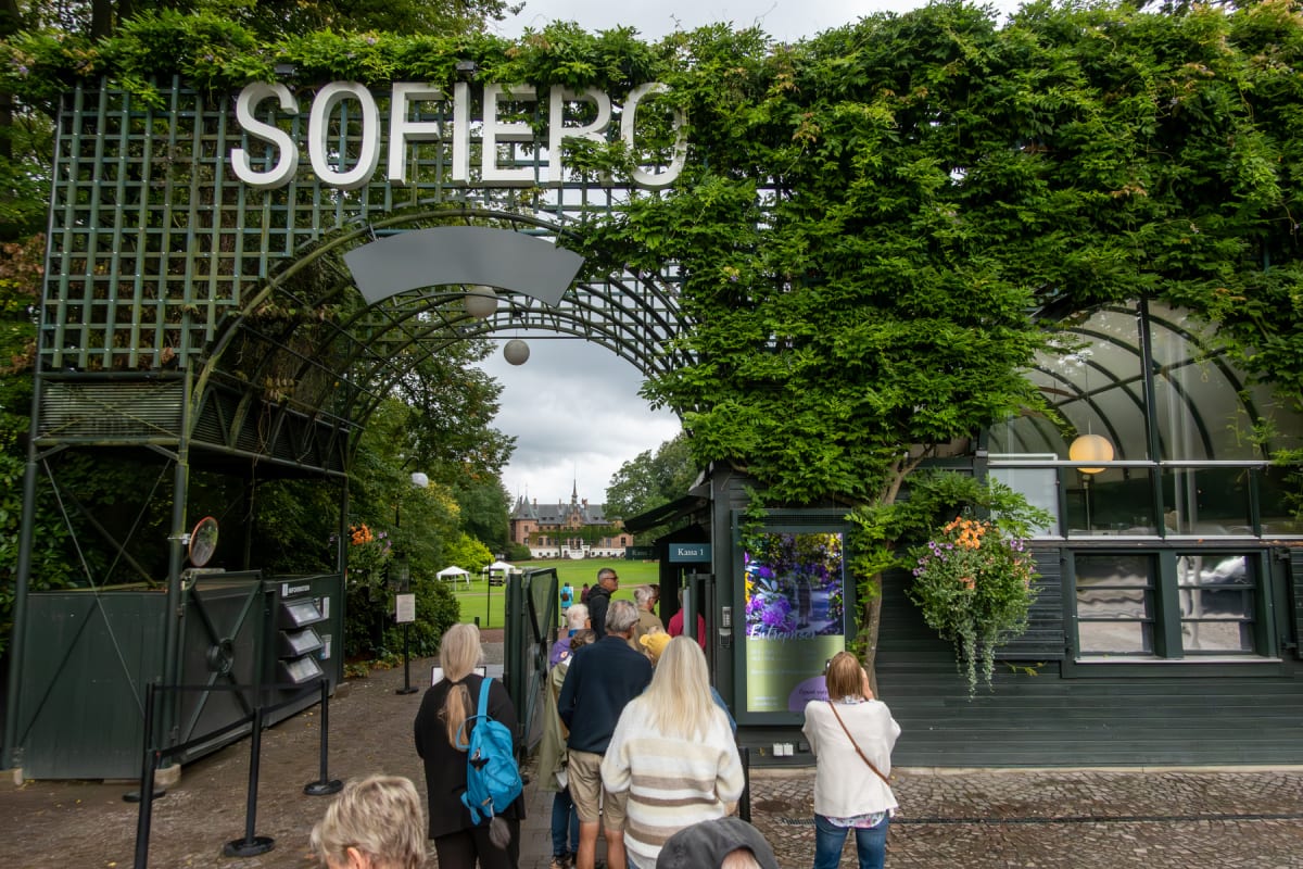 En grupp människor står i kö vid en entrégrind med en stor båge täckt av grön växtlighet. Ovanför bågen står texten "SOFIERO". Bredvid kön finns en byggnad, delvis täckt av grönska, med fönster och en digital skylt. I bakgrunden syns ett slott och en