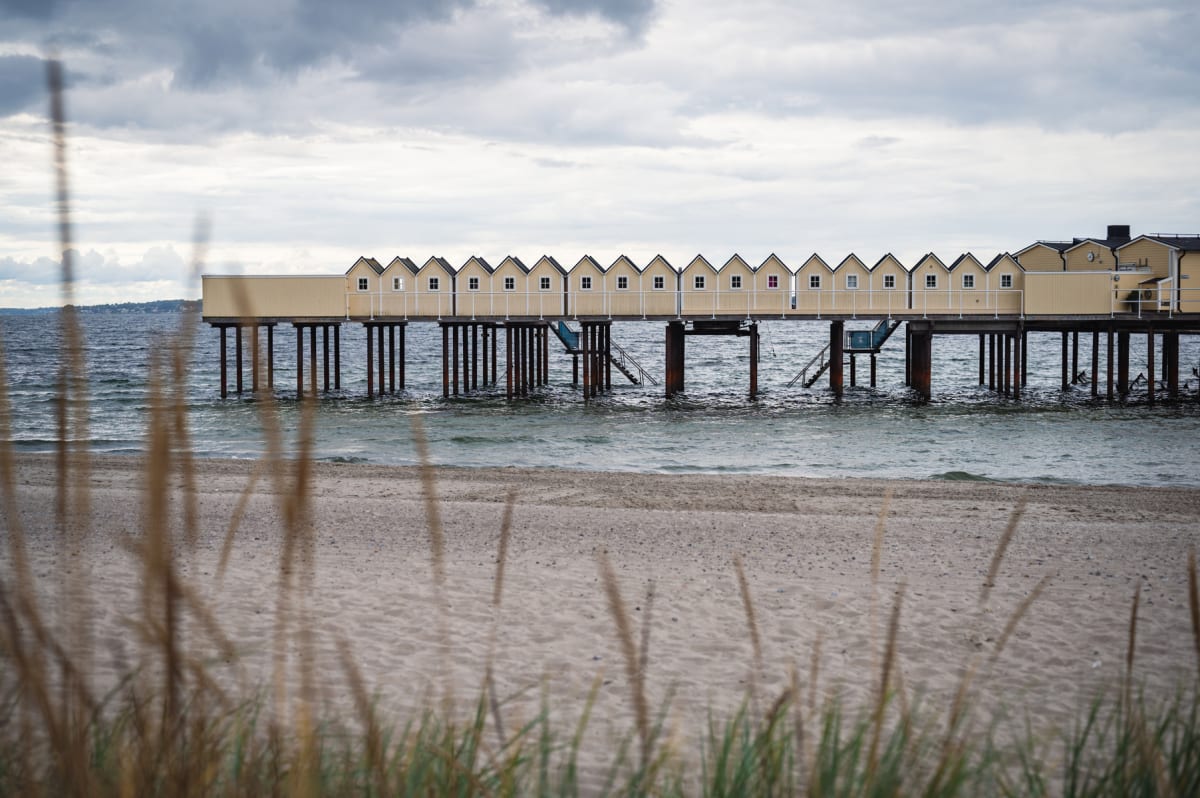 En sandstrand i förgrunden med grässtrån. I mellangrunden syns en havsutsikt med ett långsträckt, gult kallbadhus på pålar ovanför vattnet. Kallbadhuset har flera små fönster och spetsiga tak. En gråmulen himmel täcker bakgrunden.