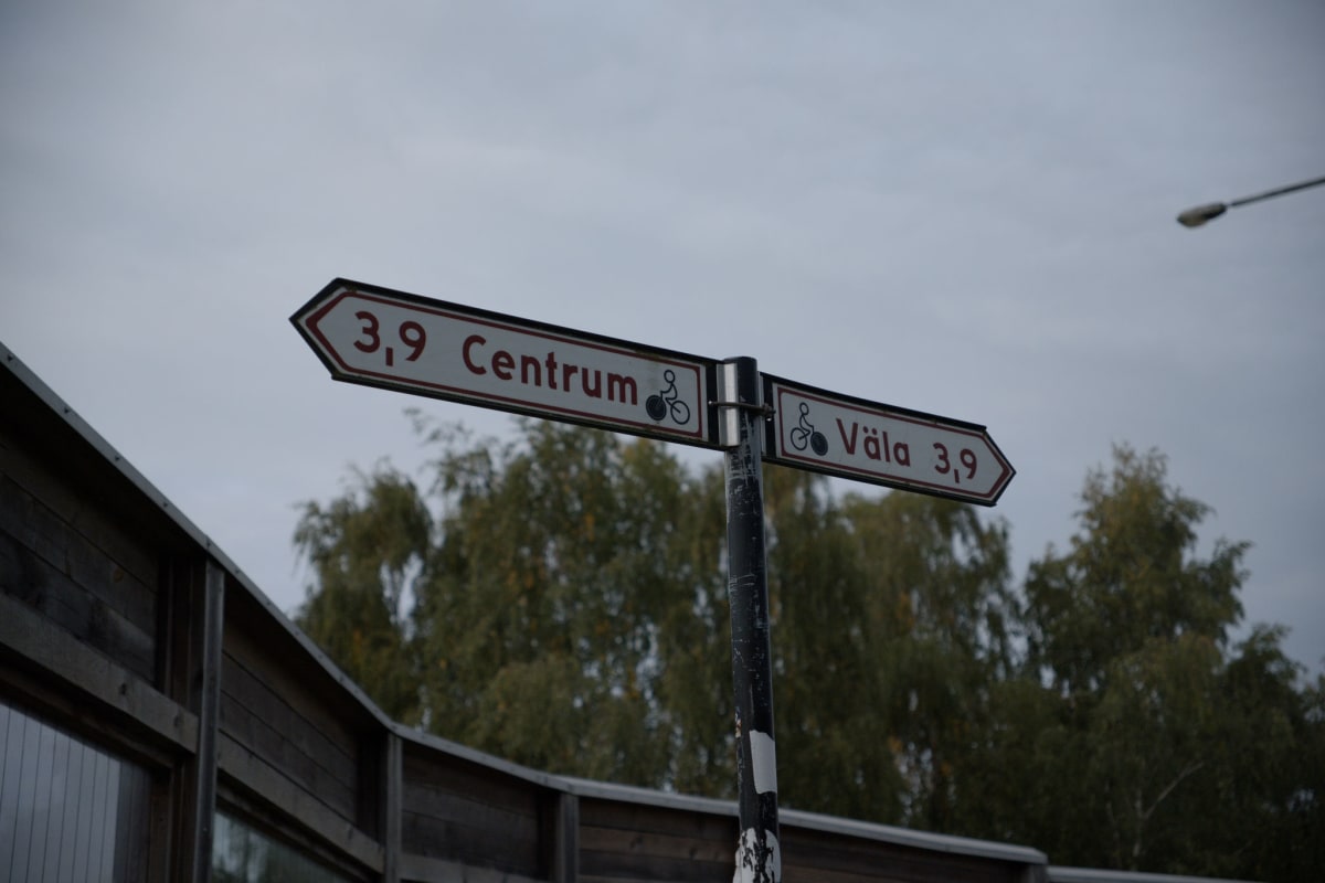 En vägvisare med två skyltar pekar ut riktningarna mot "Centrum" och "Väla", båda 3,9 enheter bort, med cykelsymboler. Bakom stolpen syns gröna lövträd och en mörk trästruktur. Himlen är mulen och grå.