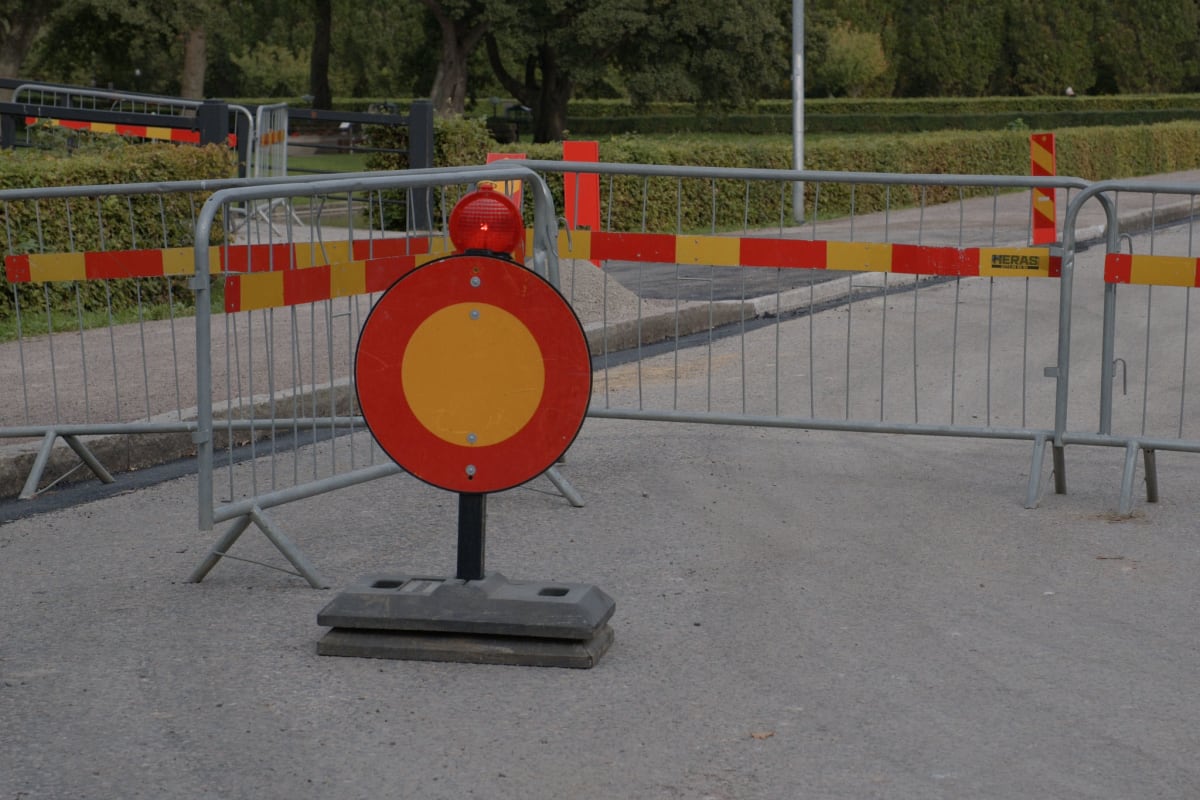 En väg är avspärrad med två metallstaket och en rund röd och gul förbudsskylt med en blinkande röd varningslampa. Skylten står på marken framför staketen. I bakgrunden syns grönska som buskar och träd under en molnig himmel.