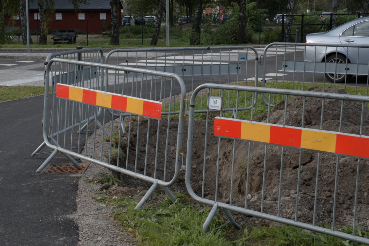 Bilden visar byggstängsel med röd-gul randig markering som avgränsar ett område med uppgrävd jord och gräs från en asfalterad väg. I bakgrunden syns träd, en röd byggnad och del av en silverfärgad personbil.