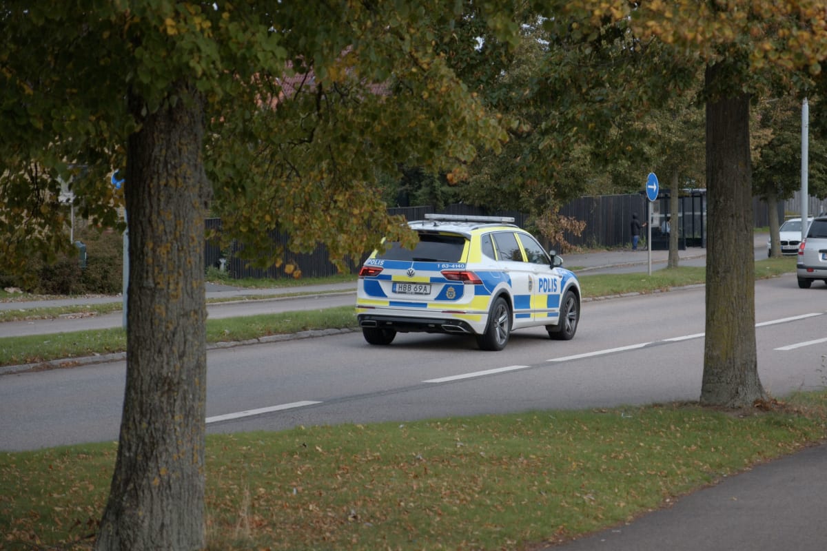 En svensk polisfordon, en vit SUV med blå och gula ränder samt texten "POLIS", kör på en asfalterad väg. Träd med löv som börjar ändra färg kantar vägen, och en gräsremsa syns bredvid vägbanan. En person står vid en busshållplats i bakgrunden.