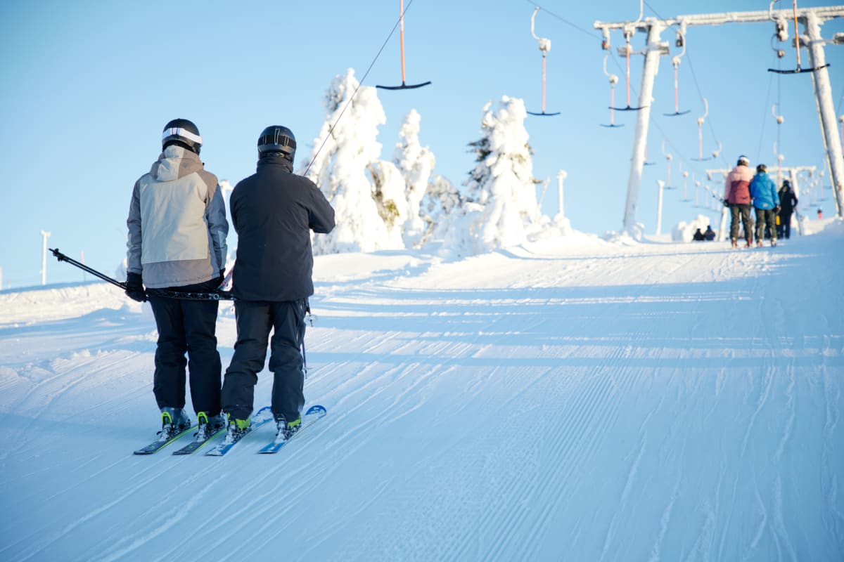 Två personer i skidutrustning syns bakifrån, stående på skidor på en snötäckt backe under en klarblå himmel. De är på väg mot en T-lift som är synlig i bakgrunden. Ett antal snötäckta träd och fler personer på skidor syns längre bort. Filnamnet indik