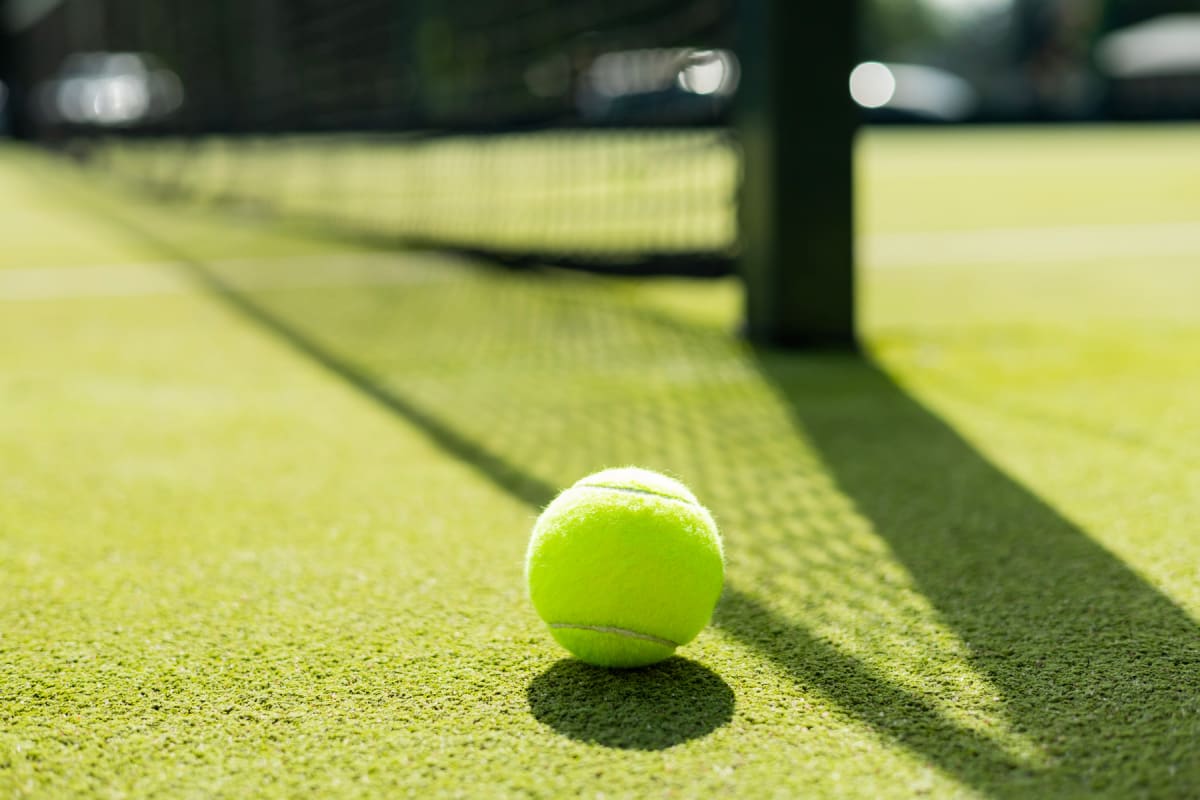 En ljusgrön tennisboll ligger på en konstgräsplan för tennis eller padel med en tydlig skugga bredvid. I bakgrunden syns ett nät och stolpar, delvis suddiga.
