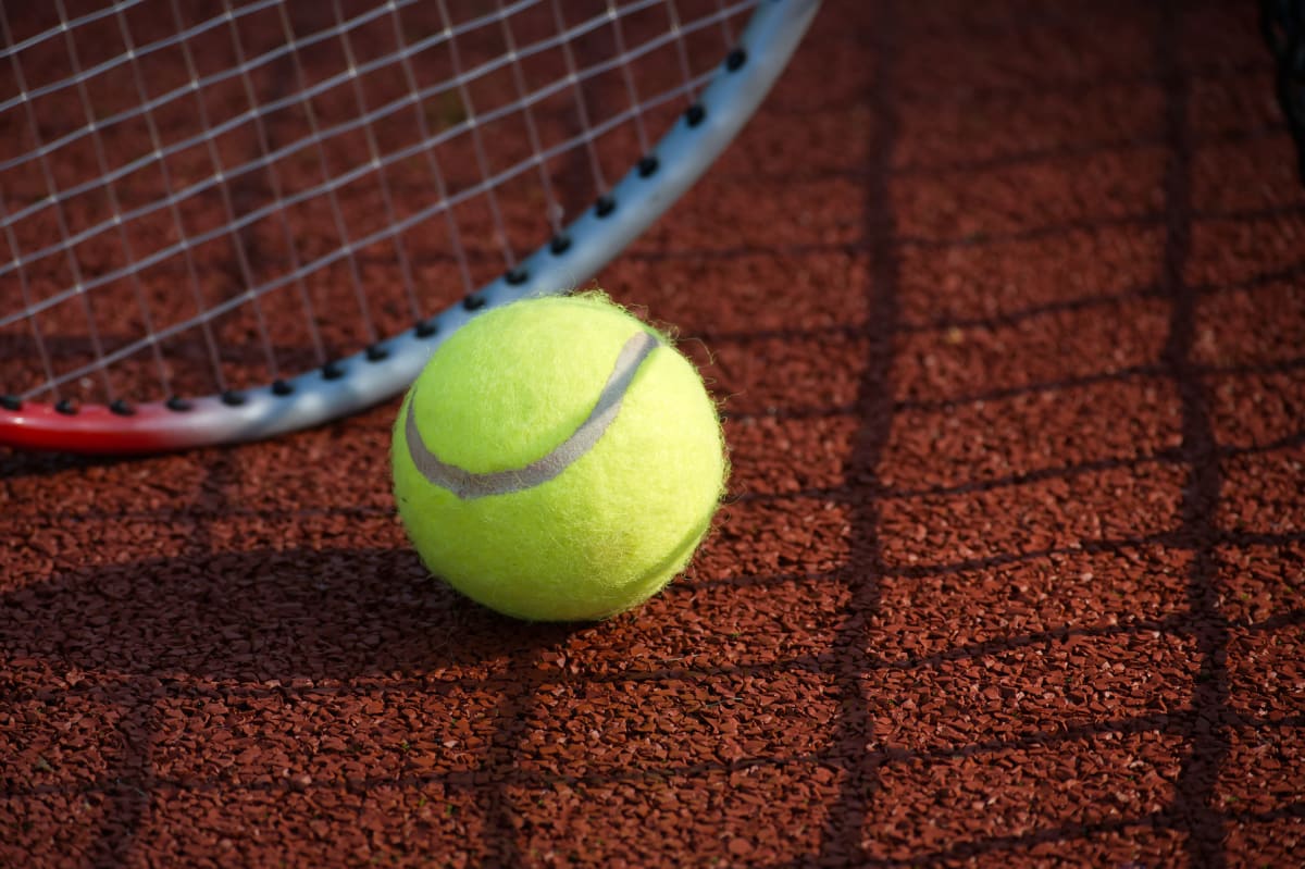 En gul tennisboll med en grå rand ligger på en rödbrun "clay court"-liknande yta. Bakom bollen syns en del av ett tennisracket med röd och silverfärgad ram. Ytan har en tydlig skugga som liknar ett tennisnät.