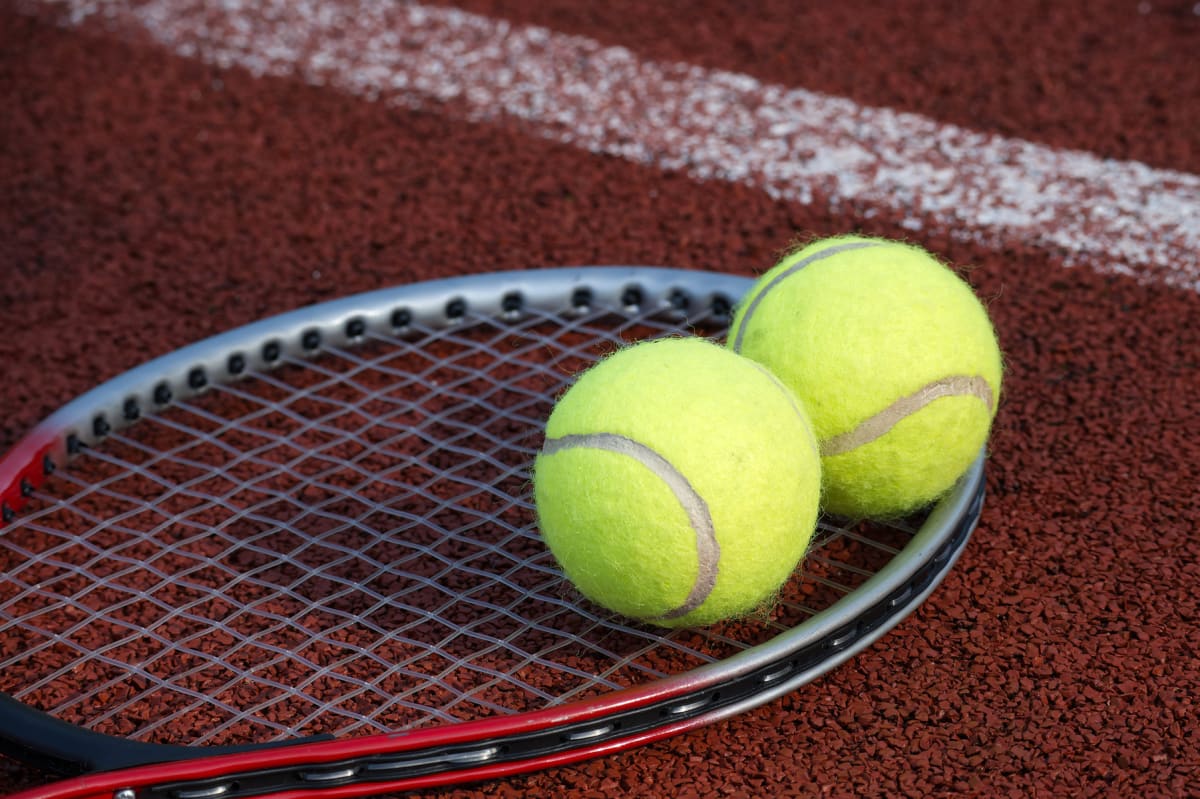 Två gröngula tennisbollar vilar på strängarna av ett tennisracket, som ligger på en rödbrun tennisbana. En vit linje syns i bakgrunden på tennisbanan.