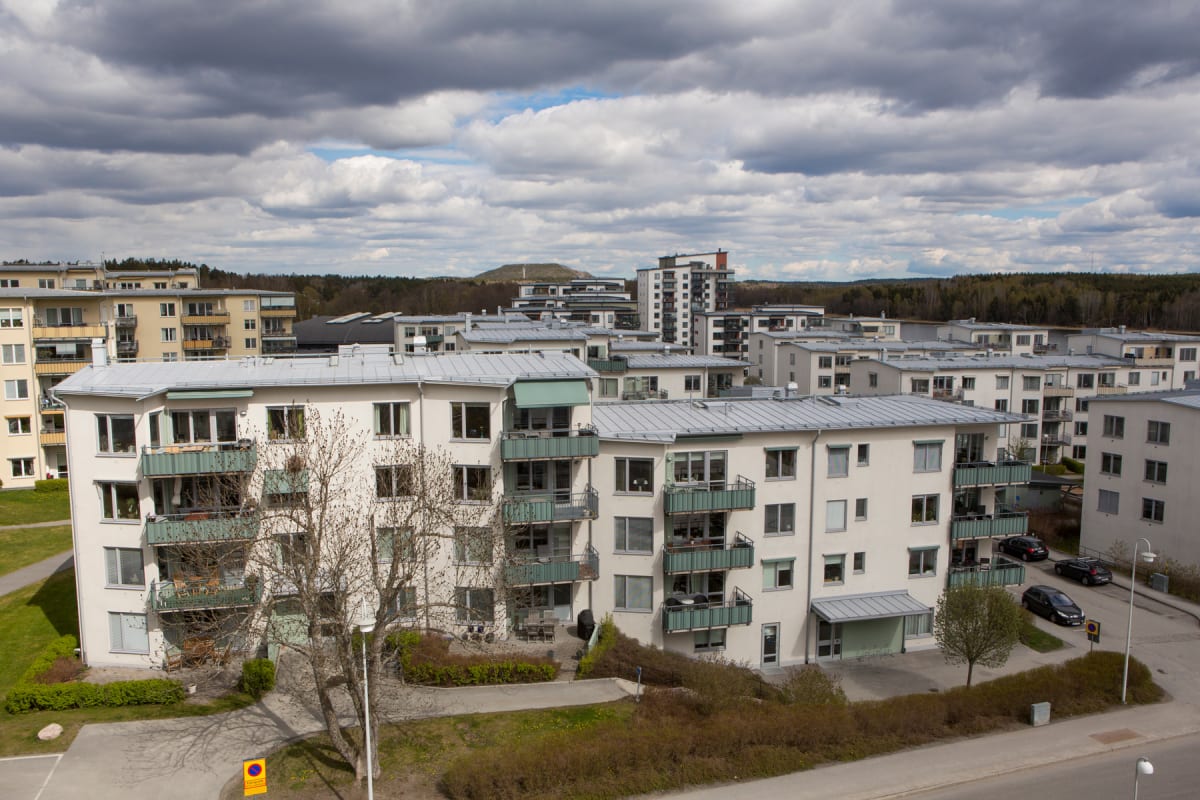 En vidvinkelbild som visar en översikt av ett bostadsområde i Hägernäs, Täby, Sverige. Flera flerfamiljshus med ljusa fasader och balkonger syns under en molnig himmel. I förgrunden och mellan byggnaderna finns grönområden med gräs, buskar och träd s