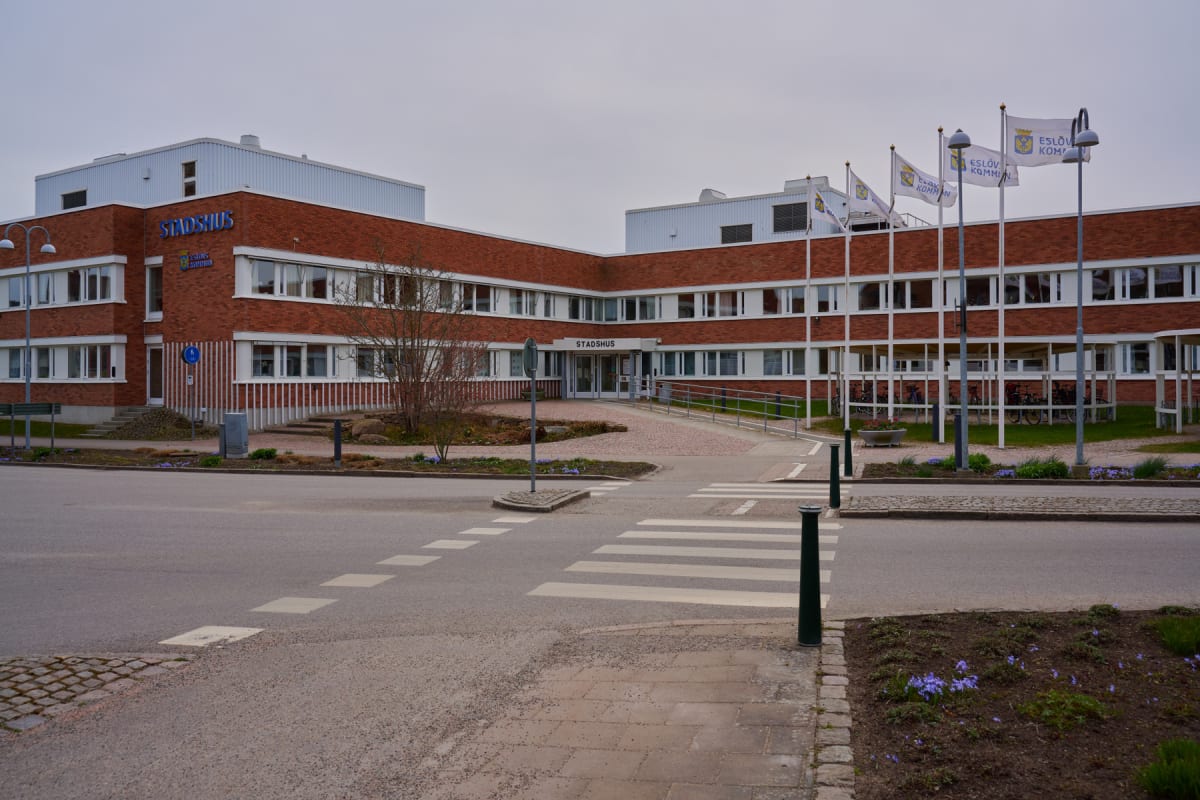 En röd tegelbyggnad med texten "STADSHUS" i blått över huvudentrén och logotypen "ESLÖV KOMMUN" syns under texten "STADSHUS" på byggnaden. Framför byggnaden finns en asfalterad väg med ett övergångsställe och flera flaggstänger med flaggor med texten