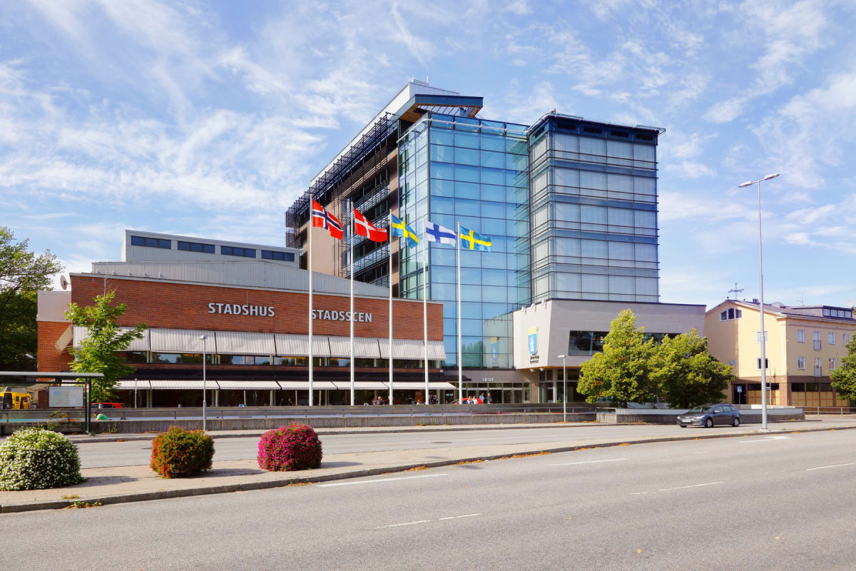 En fasadvy av Södertälje stadshus, ett byggnadskomplex med en äldre röd tegelbyggnad märkt "STADSHUS" och "STADSSCEN" och en modernare del med blåglasfasad. Framför byggnaden syns flaggstänger med flaggor från Norge, Danmark, Sverige, Finland och ytt