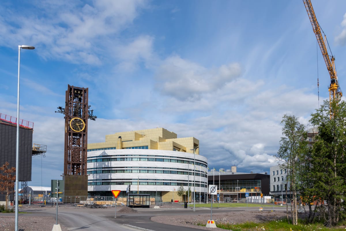En gatuscen som visar Kirunas nya stadshus med dess karakteristiska guldfärgade överdel och vita, runda byggnad, tillsammans med det gamla klocktornet i förgrunden till vänster. I bakgrunden syns en byggarbetsplats med en stor gul byggkran och flera