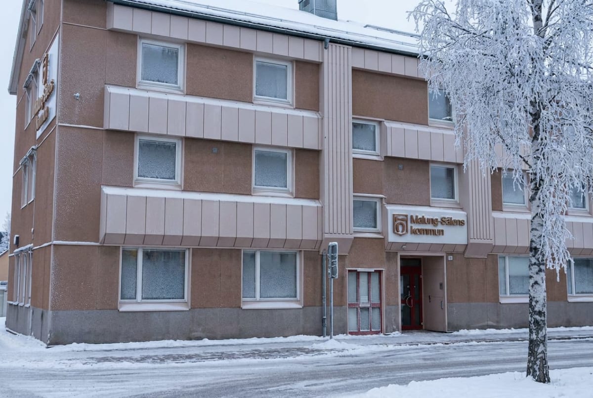 En beige och ljusbrun kommunal byggnad med frostiga fönster och en röd entrédörr, sedd från gatan en vinterdag. På fasaden finns en skylt med texten "Malung-Sälens kommun". Ett snötäckt träd står till höger om byggnaden.