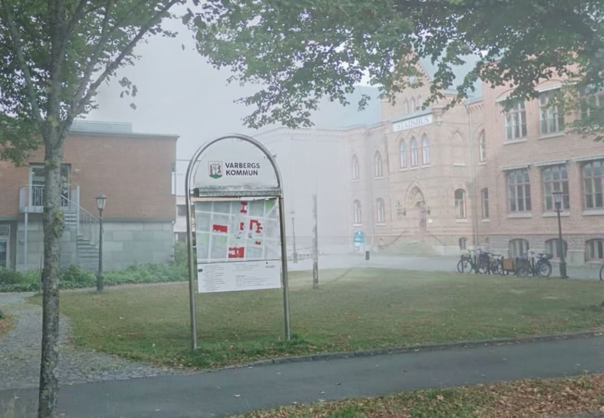 En bild som visar Varbergs Rådhus (Stadshus) och en informationsskylt för Varbergs kommun i dimmigt väder. I förgrunden syns gräs, en gångväg och ett träd med gröna löv. Flera cyklar står parkerade framför rådhuset som delvis skyms av dimman.