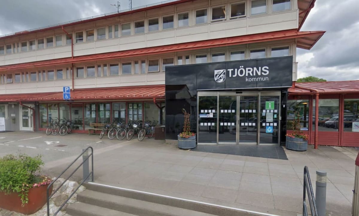 En exteriörbild av Tjörns kommunhus med ett "TJÖRNS kommun"-skylt ovanför de stora glasdörrarna. Byggnaden har flera våningar med fönster och röda tak. Framför ingången står flera cyklar parkerade, och det finns blomkrukor samt en parkeringsskylt för