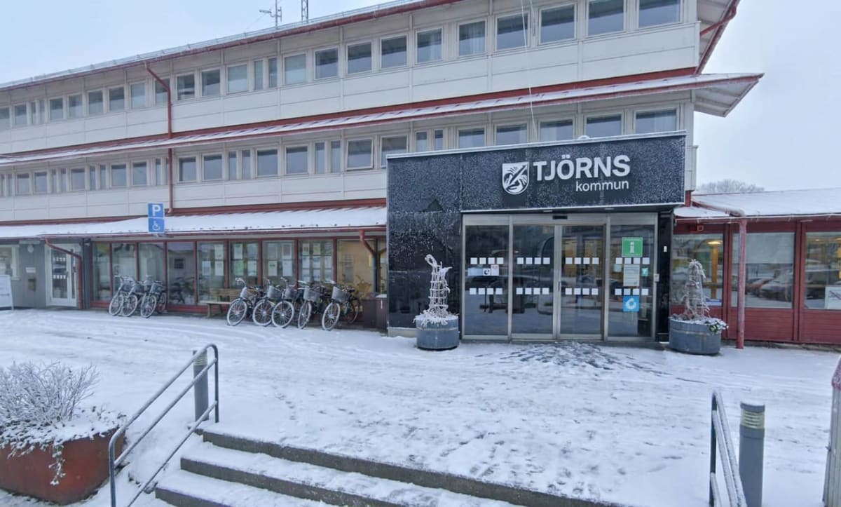En exteriörbild av Tjörns kommunhus med snö på marken framför byggnaden. Fasaden är beige och rödbrun, med många fönster. Entrén har en svart skylt med texten "TJÖRNS kommun" och glasdörrar. Flera cyklar står parkerade längs fasaden. En trappa med rä