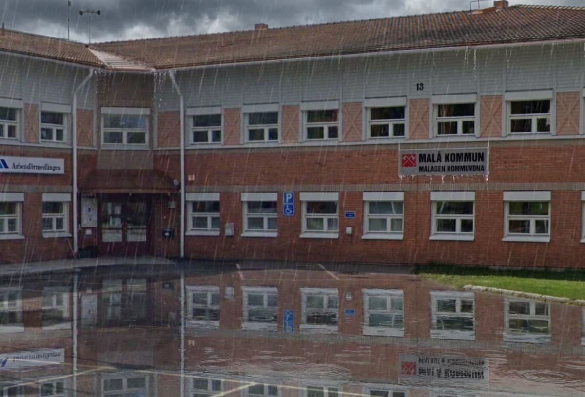 En röd tegelbyggnad med många fönster syns under regnigt väder. På byggnaden finns skyltar för "Arbetsförmedlingen" och "Malå Kommun". En stor vattenpöl på marken reflekterar byggnaden, och regndroppar faller.