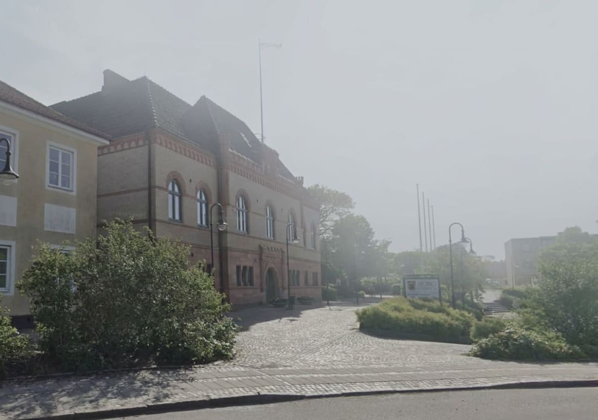 En ljus exteriörbild av en byggnad i tegel och puts i soligt väder. Byggnaden har ett tegeltak och flera fönster, både rektangulära och välvda. Framför byggnaden finns buskar, gatsten och en gatulykta. Längre bort syns ytterligare en byggnad och flag