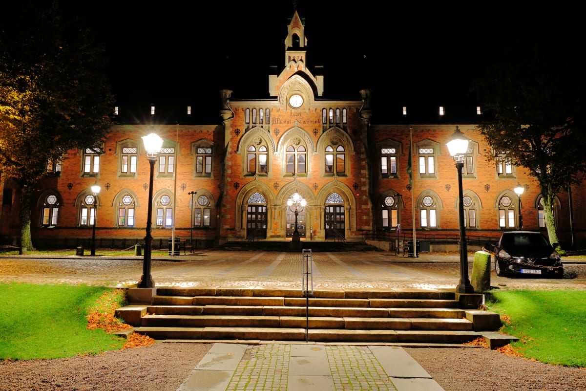 En fasadvy av en stor tegelbyggnad upplyst på kvällen. Byggnaden har många fönster med ljus, utsmyckade valv och en central klocka. Framför finns en stentrappa, gräsmattor, grusgångar och gatlyktor som lyser upp scenen. Ett träd med höstfärgade löv s