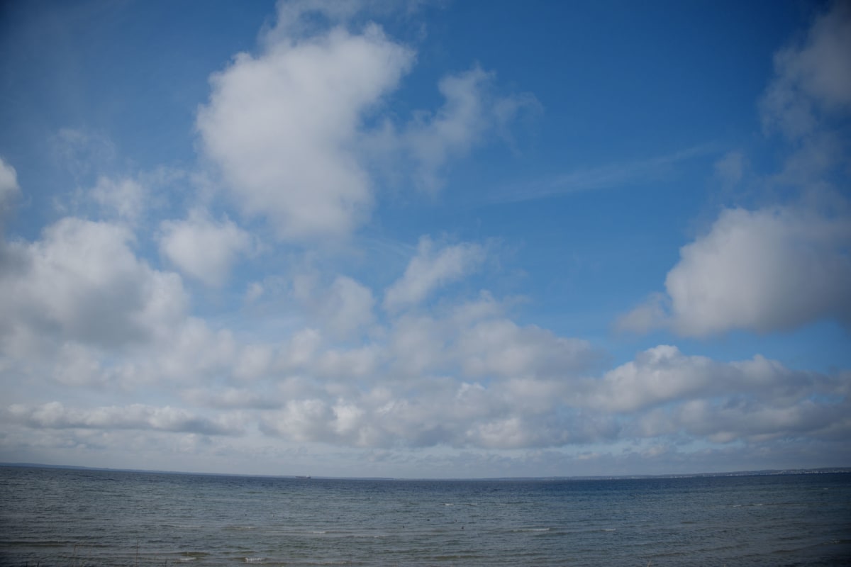 En vidsträckt vy över havet mot en horisontlinje under en blå himmel med spridda vita moln. Ett fartyg syns svagt i fjärran vid horisonten.
