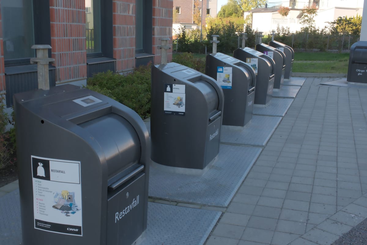 En rad grå avfalls- och återvinningsbehållare för sopsortering utomhus intill en byggnad i rött tegel. Behållarna är märkta för bland annat restavfall och tidningar.