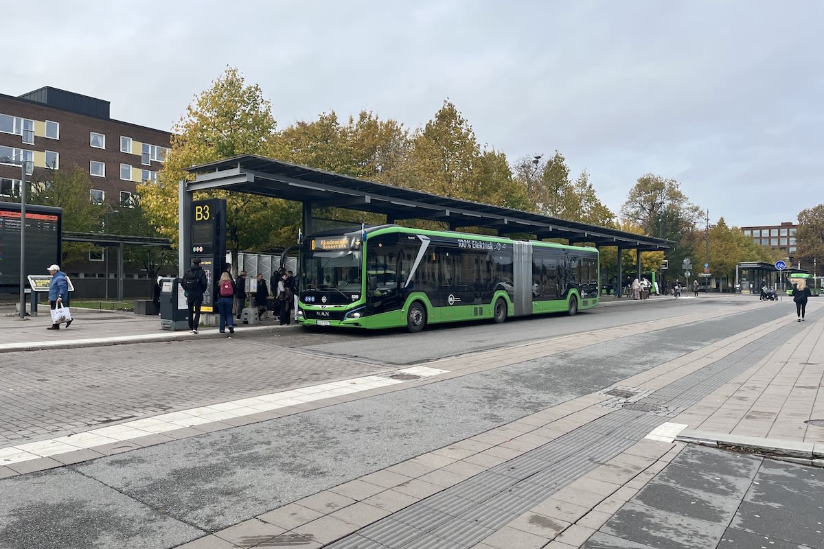 En grön eldriven ledbuss från UL står vid hållplats B3 på ett resecentrum eller busstorg med väntande passagerare under tak en höstdag.