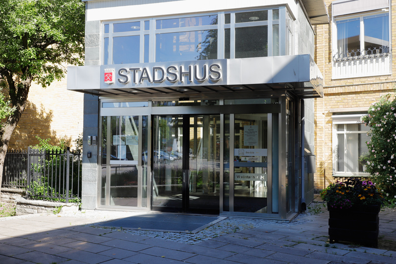 En fasadbild av en byggnad med en entré märkt "STADSHUS". Entrén har en glaskonstruktion med automatiska skjutdörrar. Ovanför dörrarna sitter en skylt med texten "STADSHUS" och en symbol som liknar ett stadsvapen. Till vänster om entrén syns en del a