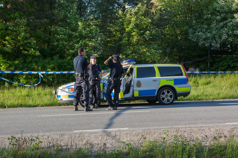 Bilden visar tre poliser och en polisfordon parkerad vid sidan av en asfaltväg. Fordonet är en vit och blå kombi med gula reflexdetaljer och texten "POLIS" synlig på sidan. Bilens framdörr är öppen. I bakgrunden syns grönskande träd och buskar. Ett v