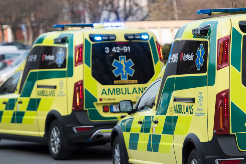Bilden visar två ambulanser parkerade utomhus. Båda ambulanserna är gula och gröna med texten "AMBULANS" och symbolen av en blå sexuddig stjärna med en orm lindad runt en stav (symbol för medicinsk kår). Den bakre ambulansen har registreringsnummer e