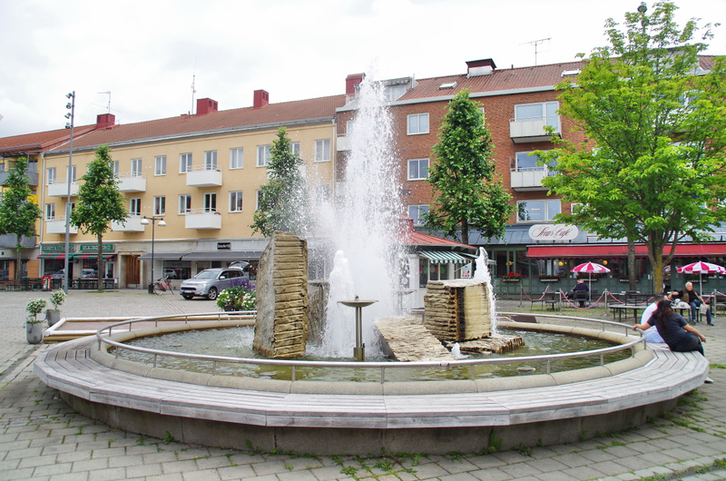 Bilden visar ett torg med en fontän i förgrunden, omgiven av en bågformad träbänk. Fontänen sprutar vatten uppåt och innehåller flera stenliknande formationer. I bakgrunden syns byggnader med mellan tre och fem våningar, där den närmaste byggnaden är