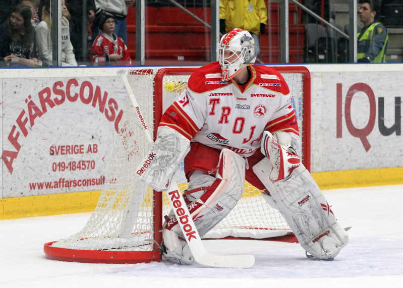 En ishockeymålvakt i vit och röd utrustning står framför ett ishockeymål på en isrink. Målvakten bär en hjälm med galler, benskydd, handskar och en målvaktsklubba. På målvaktens tröja står "TROJ" stort i rött med mindre logotyper och text som "Reebok
