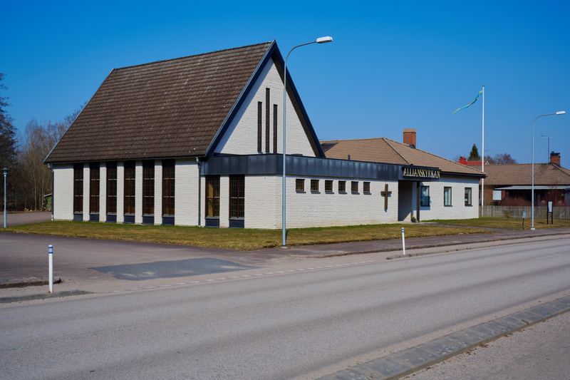 Bilden visar fasaden på en låg, vit tegelbyggnad under en klarblå himmel. Byggnaden har ett brunt tegeltak och flera fönster. Delar av byggnaden har mörkare paneler, särskilt runt fönster och vid takfoten. Marken framför byggnaden består av gräs och