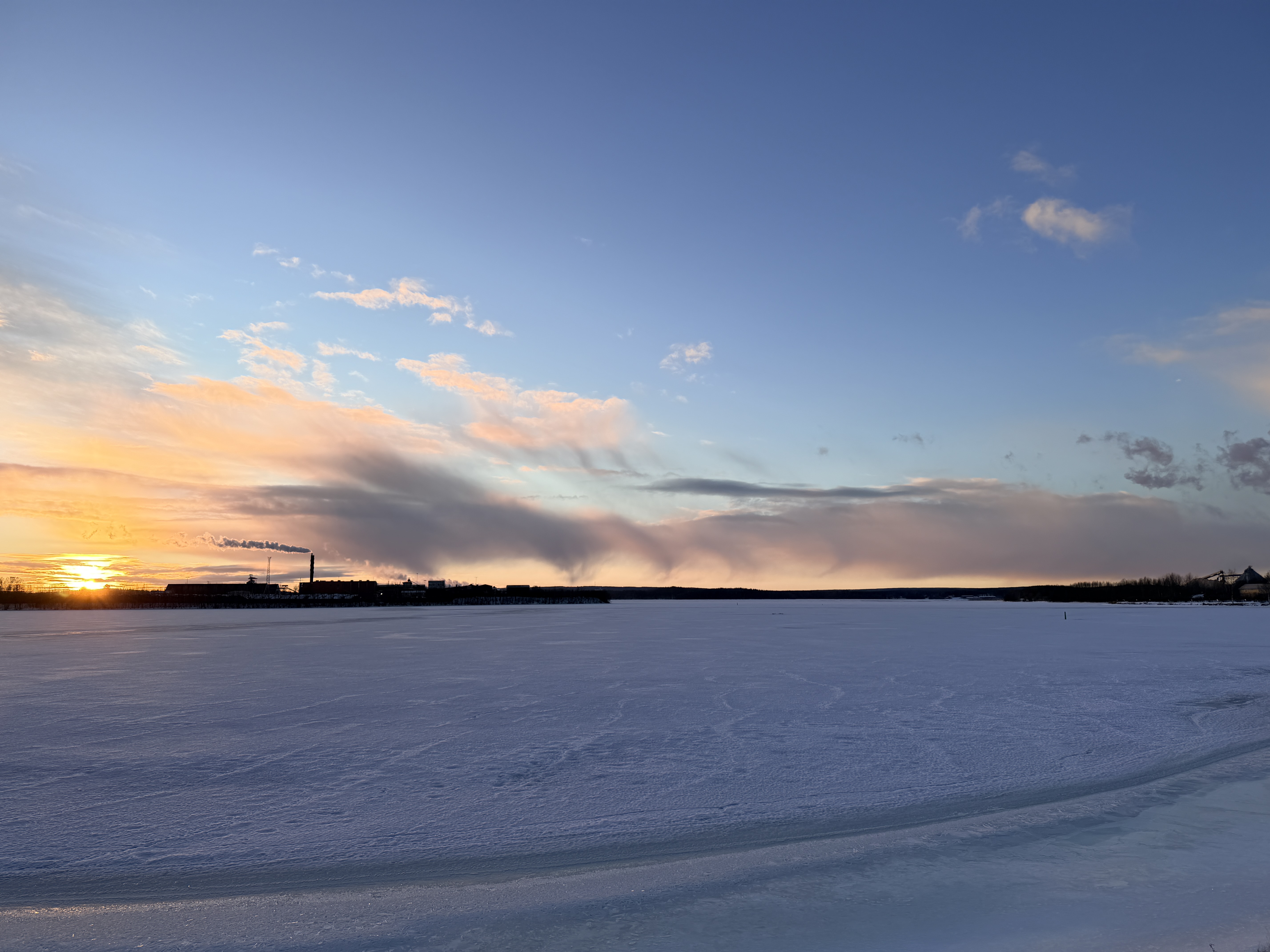 Bilden visar ett snötäckt sjö- eller havslandskap under en solnedgång eller soluppgång. Horisonten är låg, och den nedre delen av bilden domineras av is och snö med några mörkare områden som kan vara vatten eller tunn is. I bakgrunden, vid horisonten