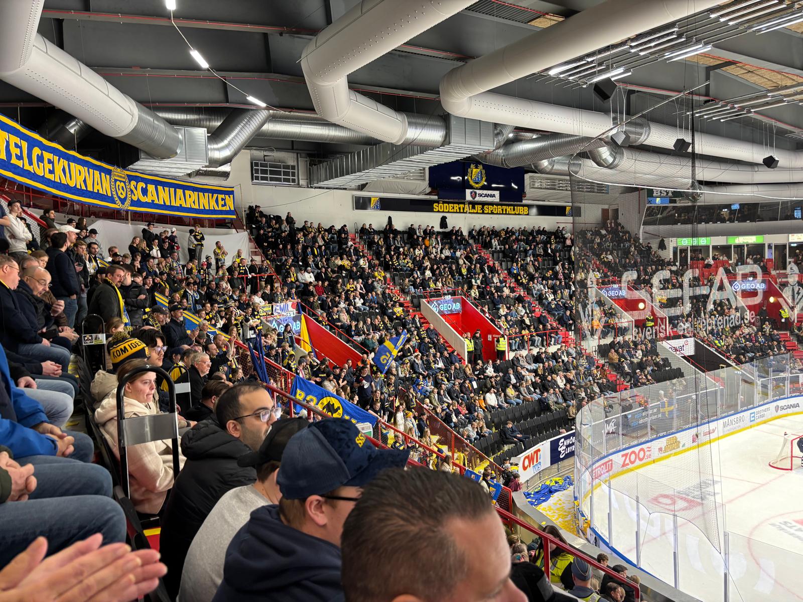 Bilden visar insidan av en ishockeyarena med publik på läktaren under en match. En stor del av läktaren är fylld med människor, varav många bär gula och svarta kläder eller accessoarer som symboliserar Södertälje Sportklubb. En del av isrinken är syn