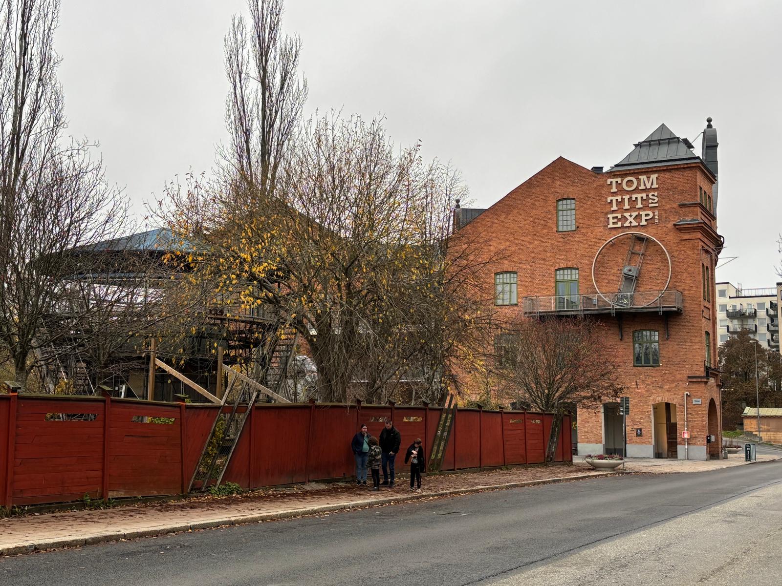En bild visar en tegelbyggnad med texten "TOM TITS EXP" vertikalt längs med fasaden. På byggnadens fasad ovanför en balkong är det monterat en stor vit cirkel med en lutande stege inuti. Träd med sparsamt med gula löv står framför byggnaden och vid s