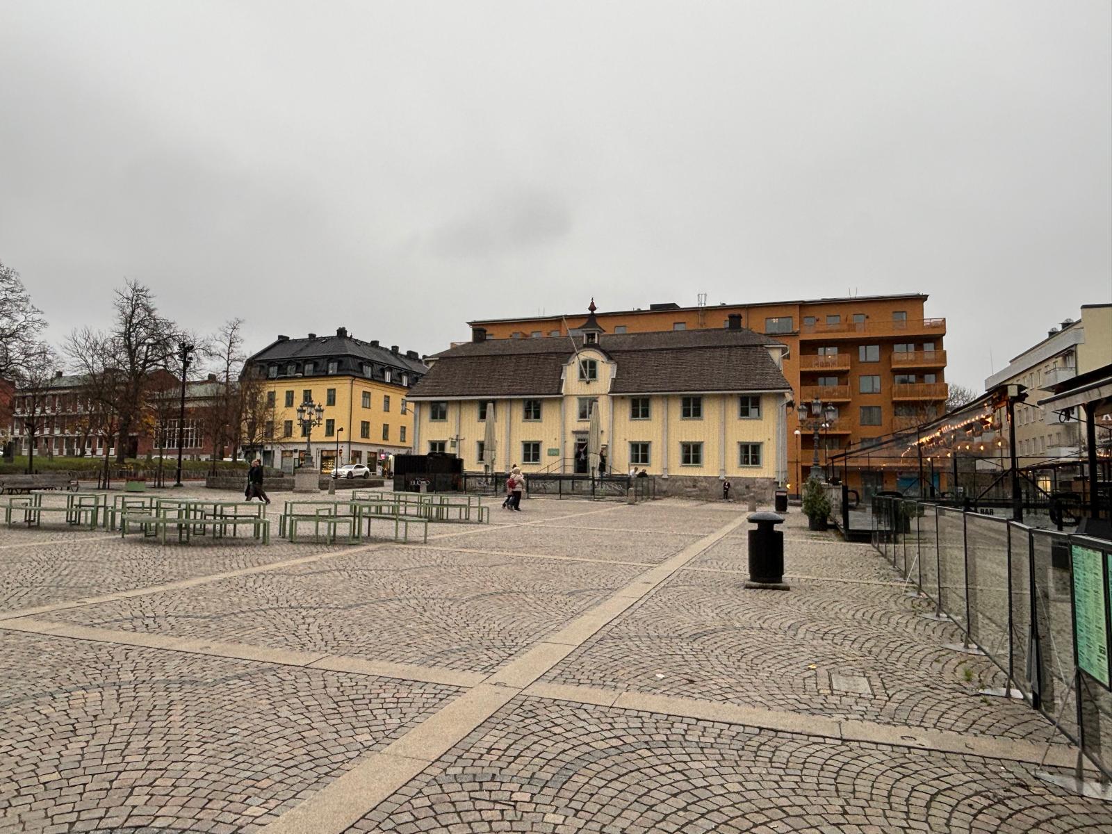 Bilden visar ett torg med kullerstenstäckning under en gråmulen himmel. I förgrunden syns tomma bord och bänkar i en ljusgrön färg. I bakgrunden finns flera byggnader, inklusive en gul tvåvåningsbyggnad med ett brunt tak som står centralt. Till vänst
