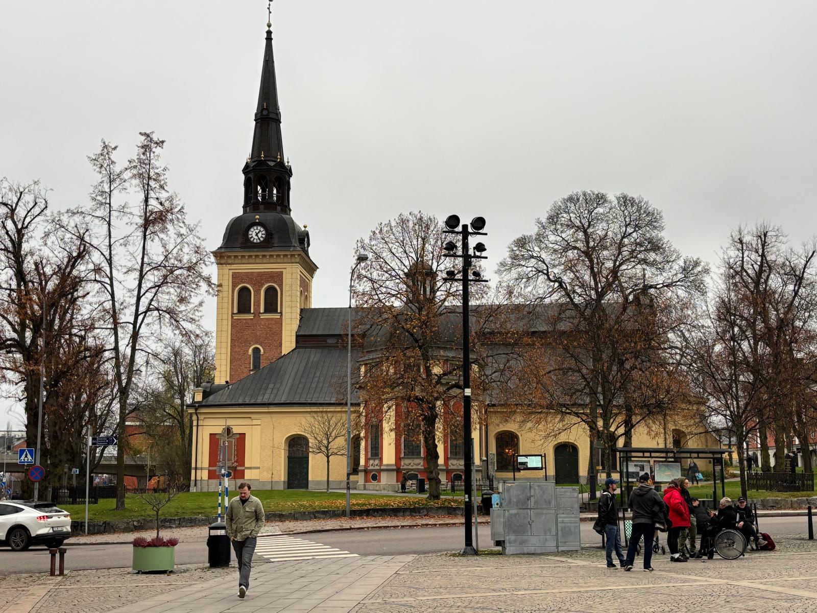 Bilden visar ett torg med en stor kyrka i bakgrunden. Kyrkan har en distinkt färgsättning med gula väggar och tegel på tornet, samt ett svart spetsigt klocktorn med urtavlor. Framför kyrkan finns flera lövträd, varav de flesta är bladlösa eller har b