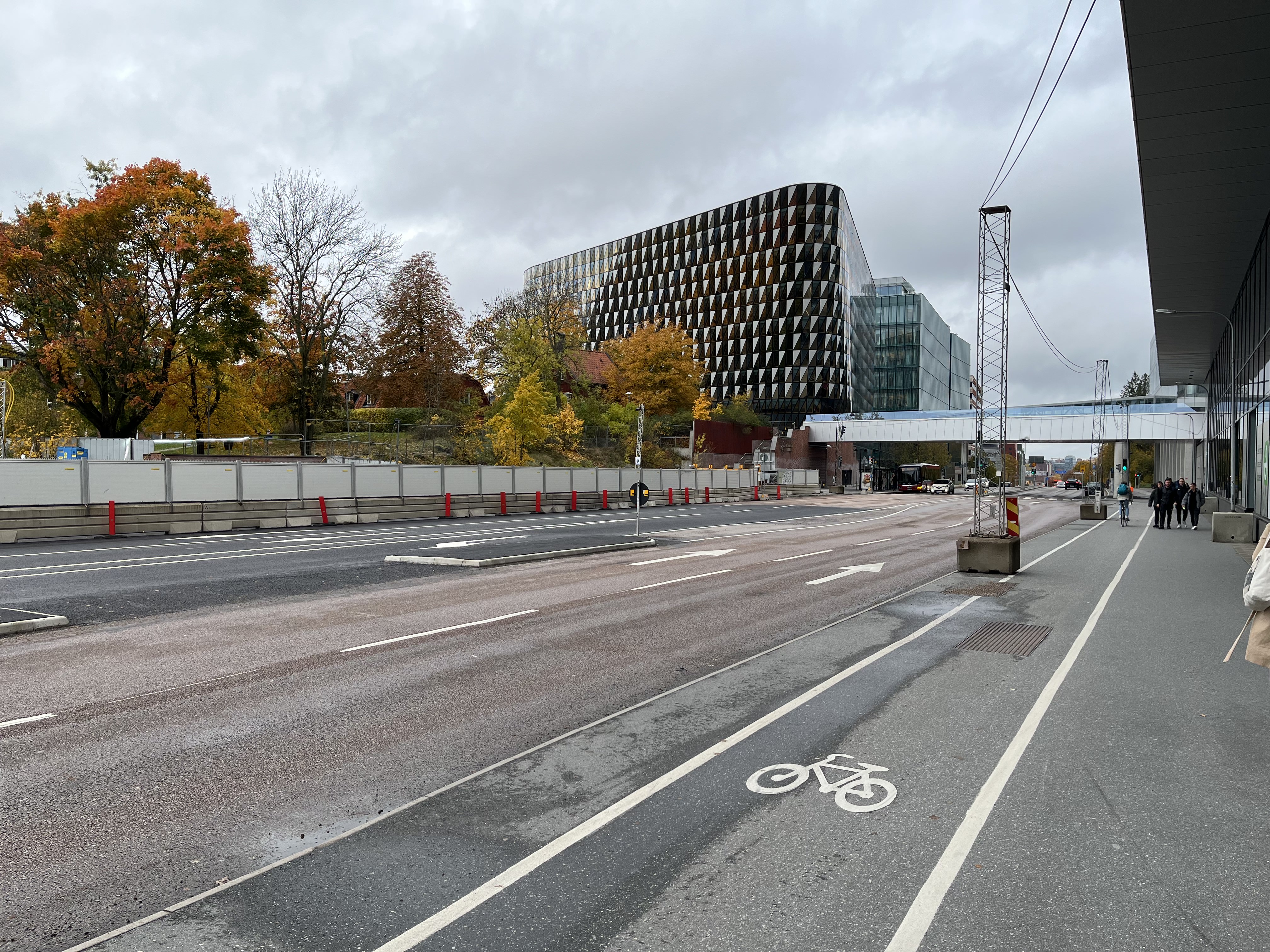 Bilden visar en stadsmiljö under dagtid med en stor väg i förgrunden som löper från vänster till höger och försvinner i bildens mitt. På vägbanan finns vita pilar och linjer. Till höger om vägen finns en cykelbana markerad med en vit cykelsymbol, bre