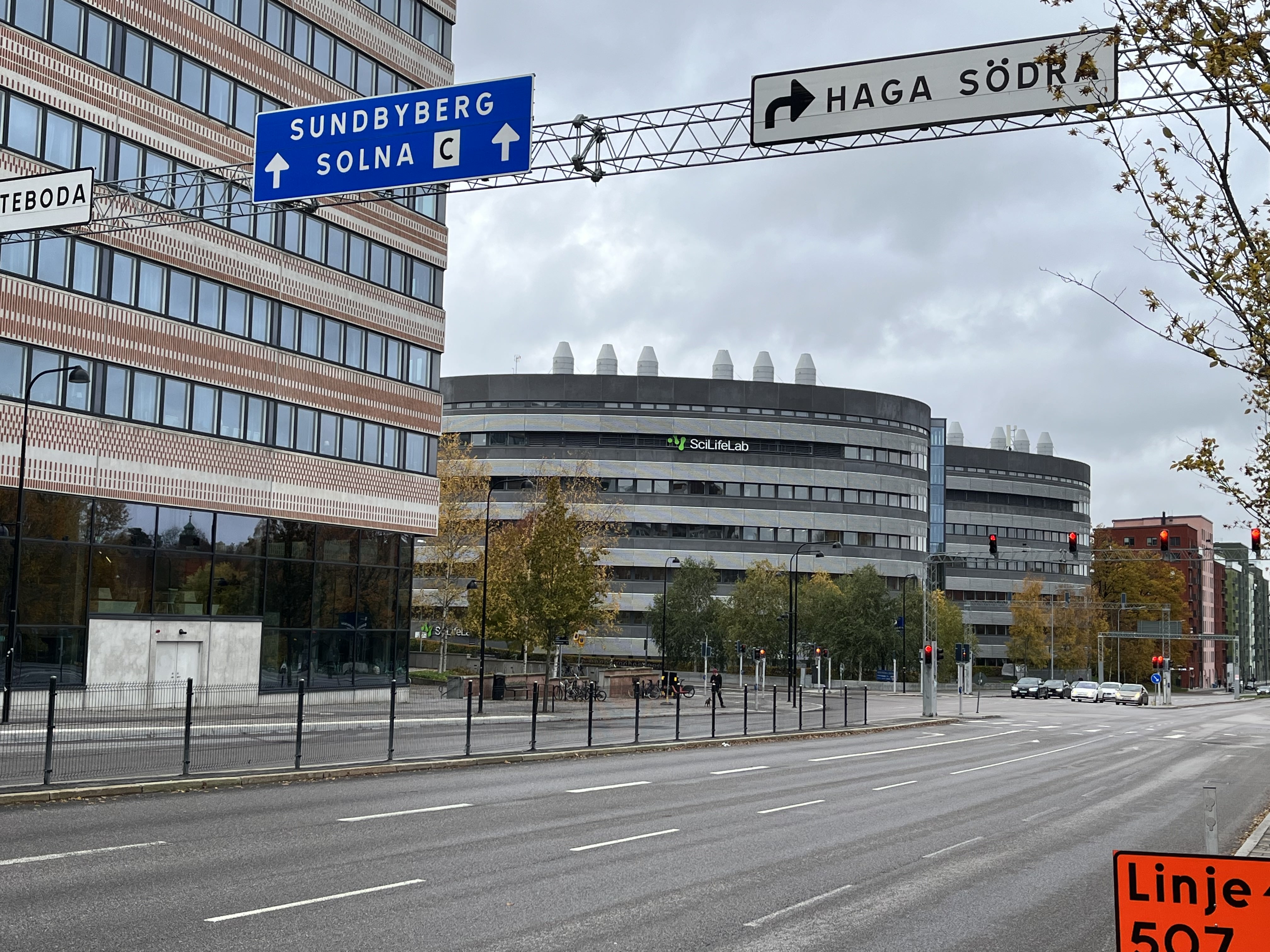En bild tagen utomhus under dagtid, föreställande en gatumiljö med byggnader och vägskyltar. I förgrunden syns en flerradig väg med vita markeringar. Längs vänster sida av vägen löper ett svart staket framför en trottoar. På vänster sida av bilden står en hög byggnad med många fönster och en mönstrad fasad i brunt och vitt. Mitt i bilden, bakom tre lövfällande träd med gulnande löv, står en stor, rundad byggnad med 'SciLifeLab' logotypen synlig på fasaden. Väggarna är grå med horisontella ränder. Trafikljus med rött sken är synliga vid flera ställen i bilden. Ovanför vägen hänger två stora vägskyltar. Den till vänster har blå bakgrund med texten 'SUNDBYBERG SOLNA C' och en pil uppåt. Den högra har vit bakgrund med texten 'HAGA SÖDRA' och en pil som pekar åt höger. I bakgrunden till höger kan man se fler byggnader i olika färger samt flera personbilar på vägen. Himlen är mulen.