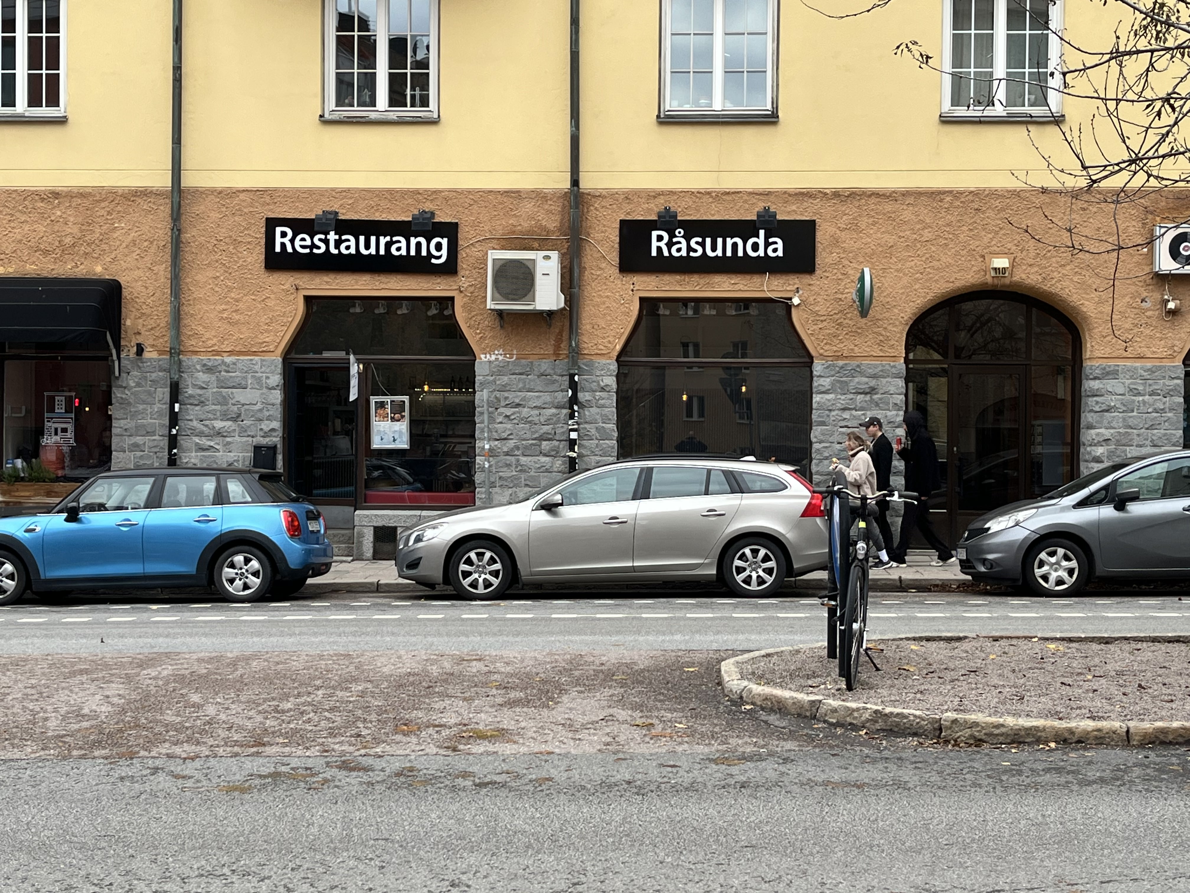 Bilden visar fasaden på en byggnad med en restaurang på markplan. Byggnaden har gul puts på övervåningen och en brunare, texturerad puts på nedervåningen som omger skyltfönster och dörrar. Ovanför två av skyltfönstren sitter svarta skyltar med vit te