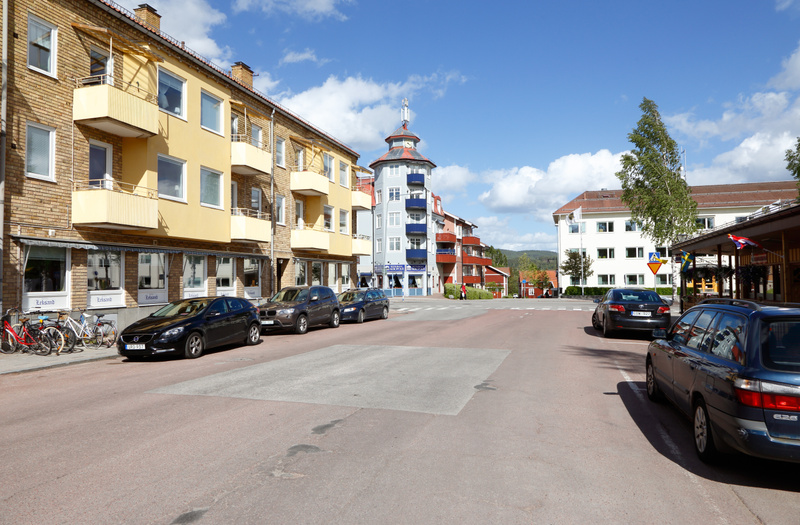 En ganska bred stadsgata syns i bildens förgrund, målad i en rödaktig färgton med några grå rektangulära områden. På båda sidor av gatan står parkerade bilar, bland annat en svart Volvo V40 och en mörkbrun BMW X3 till vänster, och en mörkblå kombibil