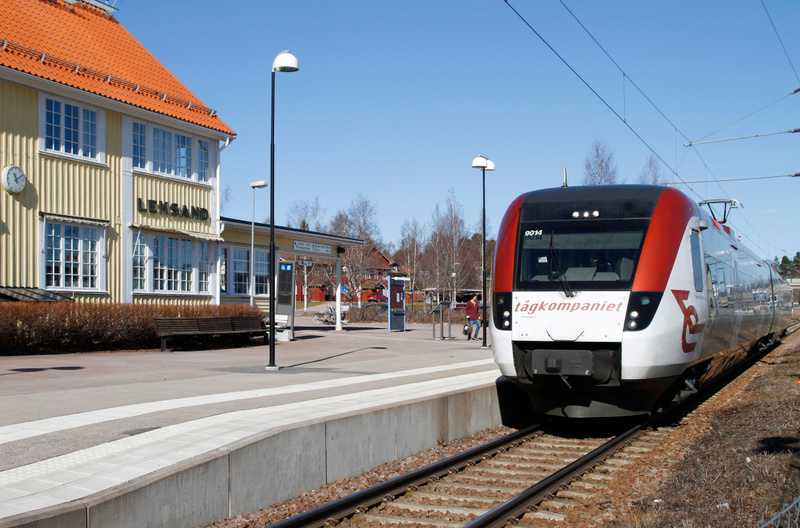 En bild tagen utomhus under dagtid som visar en tågstation. I förgrunden till höger står ett rött och vitt passagerartåg på spåret, med texten "tågkompaniet" synlig på sidan. Till vänster om tåget finns en perrong med en gul byggnad med orange tak i