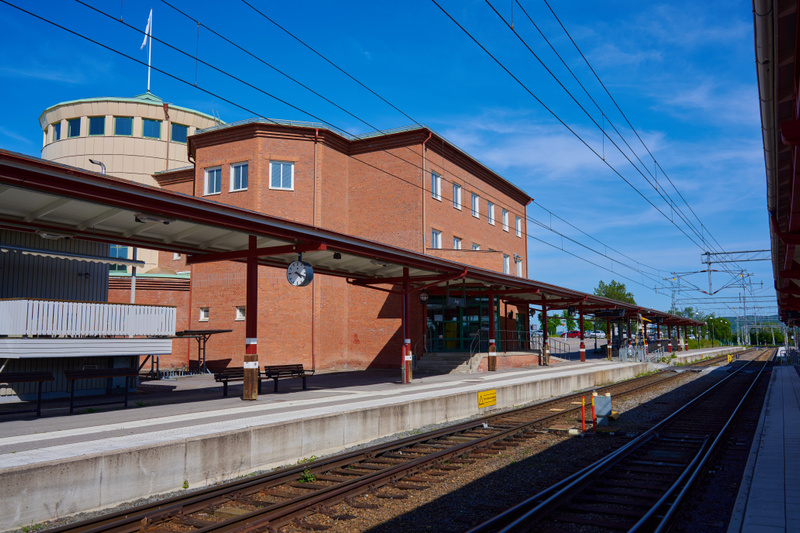 Bilden visar en järnvägsstation med spår och plattformar under klarblå himmel. Till vänster i bild syns en byggnad med tegelfasad och vita fönster, samt ett rundat torn med många fönster och en flaggstång utan flagga på toppen. Plattformarna har takk