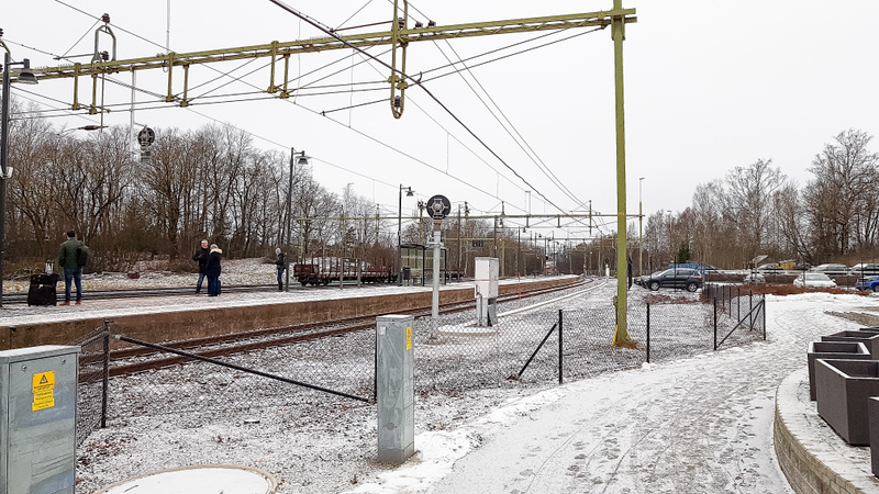 Bilden visar en järnvägsstation utomhus, troligen under vintertid med tanke på den tunna snön som täcker marken och trädens avsaknad av löv. Flera personer syns stå på perrongen. Spårområdet är snötäckt och det finns kontaktledningar och elstolpar ov