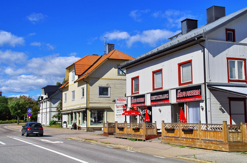Bilden visar en gata med byggnader på höger sida och ett fordon som kör på vägen på vänster sida. Två av byggnaderna har restaurangfasader med skyltar för 'Ralfs Pizzeria' och 'Restaurang'. Framför den vita byggnaden finns en uteplats med trästaket o