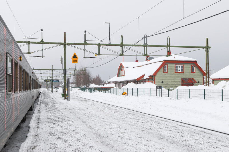 Bilden visar en järnvägsstation täckt av snö under en grå vinterdag. Ett tåg står vid en plattform till vänster, med delar av dess silverfärgade vagn synlig. Spåren är delvis snötäckta, och en plattform sträcker sig mot mitten av bilden. I bakgrunden