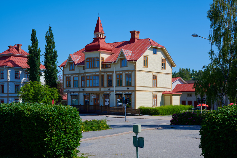 Bilden visar en gata med en stor, ljusgul trähusbyggnad med ett rött tak i centrum. Byggnaden har många fönster och en framträdande tornliknande konstruktion med spetsigt rött tak. Till vänster om huvudbyggnaden syns ett liknande, något mindre hus, o