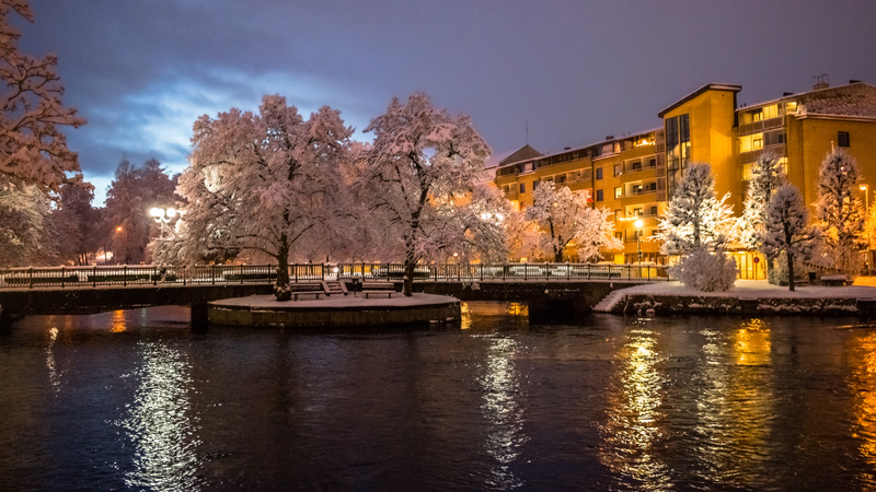 Bilden visar ett stadsmiljö på kvällen under vintern. En flod eller kanal flyter i förgrunden med mörkblått vatten som reflekterar ljusgula och vita ljus från byggnader och gatlyktor. Över vattnet sträcker sig två broar. I mitten av vattendraget finn