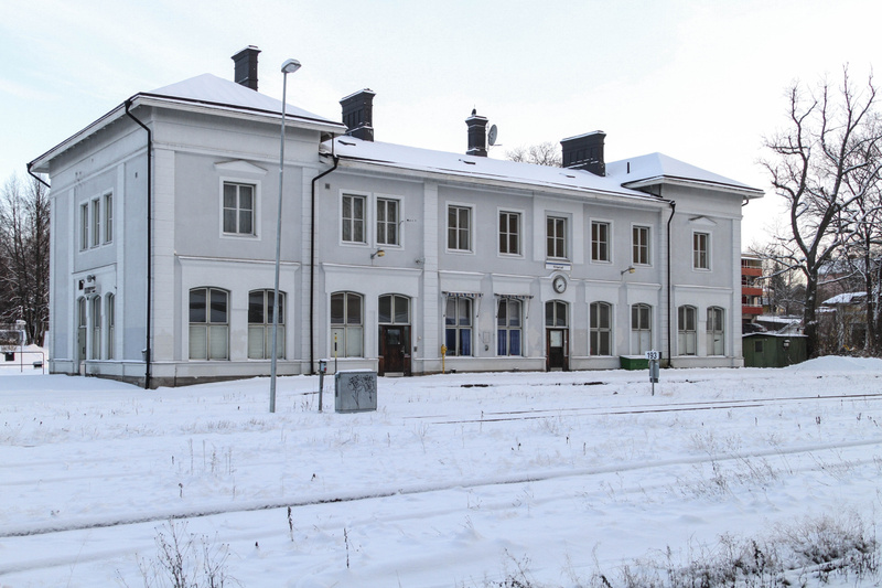 En vidvinkelbild tagen på ett snötäckt landskap med en ljusgrå byggnad i mitten av bilden. Byggnaden har flera fönster i två rader och flera dörrar längs dess framsida. Taket är också snötäckt och har en mörk skorsten. En gatlykta står framför byggna