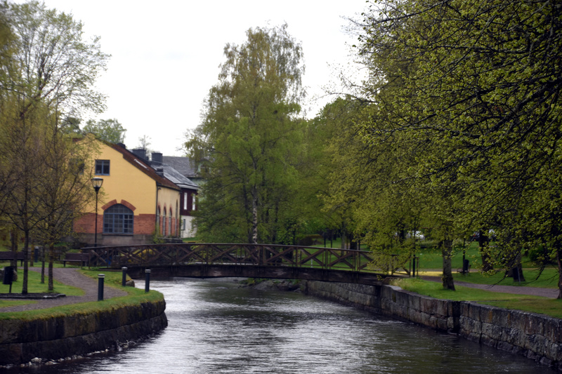 Bilden visar en kanal eller flod med mörkt vatten som flyter under en träbro. På båda sidor om vattendraget finns grönskande mark och träd med ljusgröna löv. Till vänster om kanalen syns en grusgång och ett par bänkar. Längre bak på vänster sida står