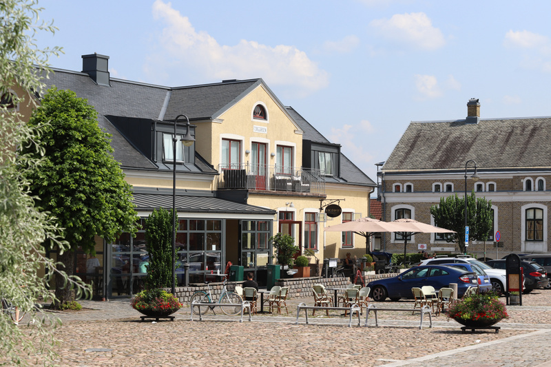 Bilden visar ett torg eller en öppen plats med kullersten i förgrunden. Till vänster syns delar av en grönskande buske eller ett träd. I mitten av bilden dominerar en gul byggnad med svart tak och flera fönster. Byggnaden har en balkong på andra våni
